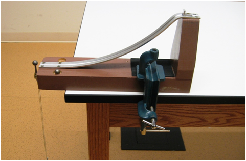 An L-shaped piece of wood is clamped down to the top of the table. A metal track is attached to the top of the vertical part of the wood and slopes down to the horizontal part of the wood to where it extends slightly off the edge of the table. A stop at the top of the track ensures balls are always launched at the same position. A steel ball sits on a stand just in front of the bottom of the track. A chain hangs down from this stand to the floor. 