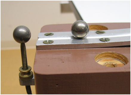 A steel ball is near the bottom edge of a horizontal metal track. Just to the left of the edge of the track is another ball sitting atop a screw. This ball is at the same height as the horizontal track.