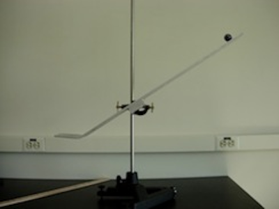 A meterstick and vertical rod are resting on a tabletop.  A metal track is attached to the rod by an adjustable clamp such that the lower end is positioned above the table. A ball is located at upper end of the track.  The lower end of the track bends into a flat section oriented parallel to the surface of the table.