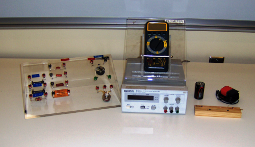 The items in the Apparatus list are displayed here. On the left, fitted on a tilted plastic stand are three columns of circuitry. The leftmost column contains resistors: from top to bottom are a 150 ohm resistor, a 700 ohm resistor, and a larger resistor not in use. The middle column contains capacitors. The rightmost column contains several light bulbs. Next to the plastic stand a digital multimeter rests within a tilted plastic stand labeled P L C meter. The multimeter sits atop a DC power supply. To the right of the power supply are a D size battery and an AC transformer with wires to connect to a DC power supply, and also a Nichrome wire and a copper wire mounted atop a small wooden block.