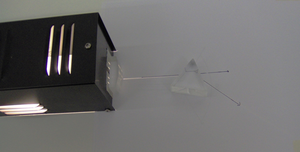 There are three objects in this figure. A plano-convex lens is placed in front of the light source to produce a single light ray. This light ray is incident on one side of the prism at some angle from the normal. The refracted ray is seen coming out of the other side of the prism. There is also a line that traces the original incident ray.