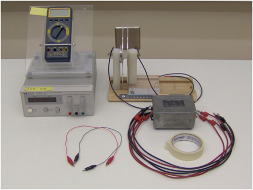 The supplies for this lab are displayed on top of a table. There are two alligator clip leads, a D C power supply, a circular parallel plate capacitor, a multimeter, an interface box, masking tape, and several banana-banana leads. 