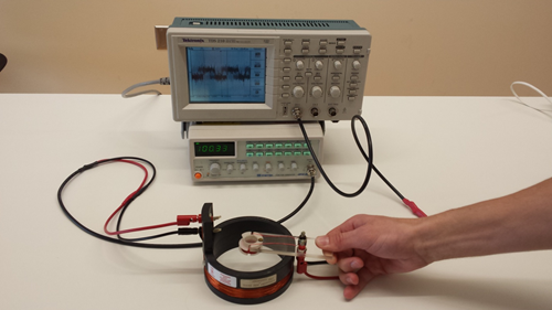 Two coils are at the center of the image.  The large coil sits atop the table and is connected by cable to the function generator.  The small coil is held above the large coil and is connected by cable to the oscilloscope.